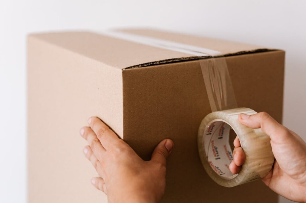 Crop faceless male sealing packed carton box with tape against white wall in daylight during relocation