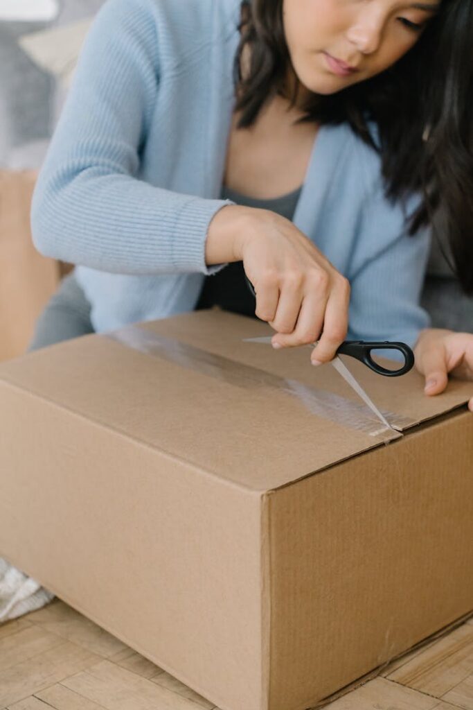 Asian woman using scissors to open a large cardboard box indoors.